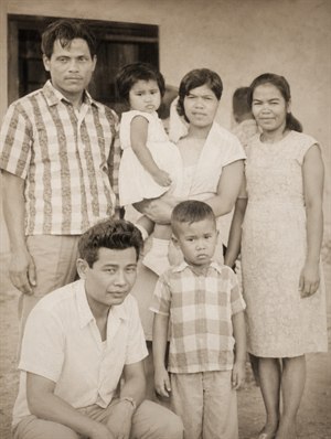Kung Fu Hand Painting - Restored sepia family portrait showing six people posed outdoors in front of a simple building wall and dark doorway. The photo has repaired cracks, softened stains, improved contrast, and clearer faces. A man stands left, a woman stands right, a central woman holds a toddler, with a young boy and crouching man in front.