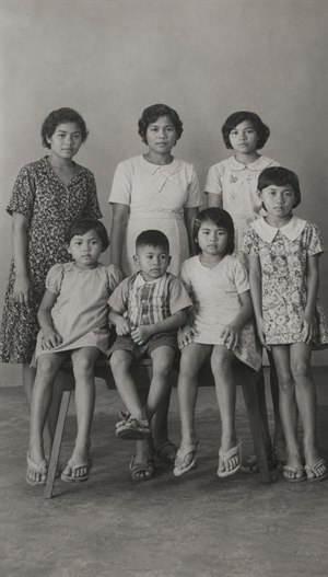 Kung Fu Hand Painting - Restored vintage black-and-white family portrait showing seven people posed formally in a simple studio setting. The cleaned image reveals clearer faces, clothing textures, sandals, bench details, and soft floor shadows. The wider framing gives the portrait more breathing room while preserving the quiet, nostalgic character of the original photograph.